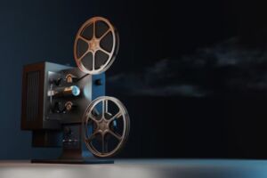 Ein alter Filmprojektor steht in einem dunklen Raum. Zwei große Filmrollen sind auf dem Projektor montiert, und ein Lichtstrahl projiziert das Bild in die Ferne. Der Hintergrund ist dunkel gehalten, und der Filmprojektor ist gut beleuchtet, was seine metallischen Details hervorhebt.
