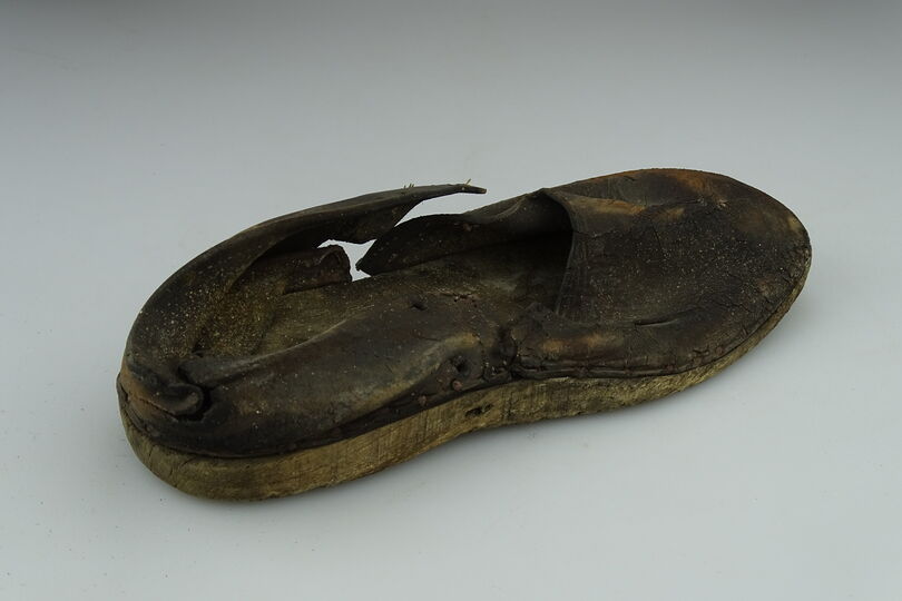 Die Abbildung zeigt einen alten Arbeitsschuh. Die Sohle besteht aus Holz. Das Obermaterial aus Leder. Der Schuh ist kaputt und dreck.