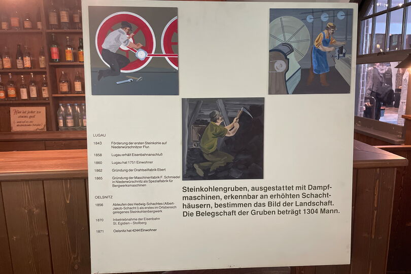 Es ist eine quadratische Tafel abgebildet. Sie hat einen hellen Hintergrund. Auf der Tafel befinden sich drei quadratische Abbildungen. Diese sind handgemalt. Das Bild oben links zeigt einen Mann, der an einem Rad einer Dampflokomotive arbeitet. Rechts oben befindet sich eine weitere Abbildung. Auf ihr sind links Teile einer Dampfmaschine zu sehen, daneben steht ein Mann, der an einer Werkbank arbeitet. Auf dem Bild in der Mitte ist ein Mann dargestellt, der in einem Bergwerk arbeitet.  Links neben diesem Bild und darunter steht Text.