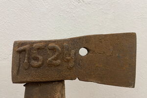 Die Abbildung zeigt einen Kaukamm. Dieser wird vor eine weiße Wand gehalten. Der Kaukamm ist ein Grubenbeil. Dieses besteht aus einem Kopf aus Metall, in dem ein Stiel aus Holz gesteckt ist. Auf dem Kopf steht die Zahl „1524“. Daneben ist ein Loch.