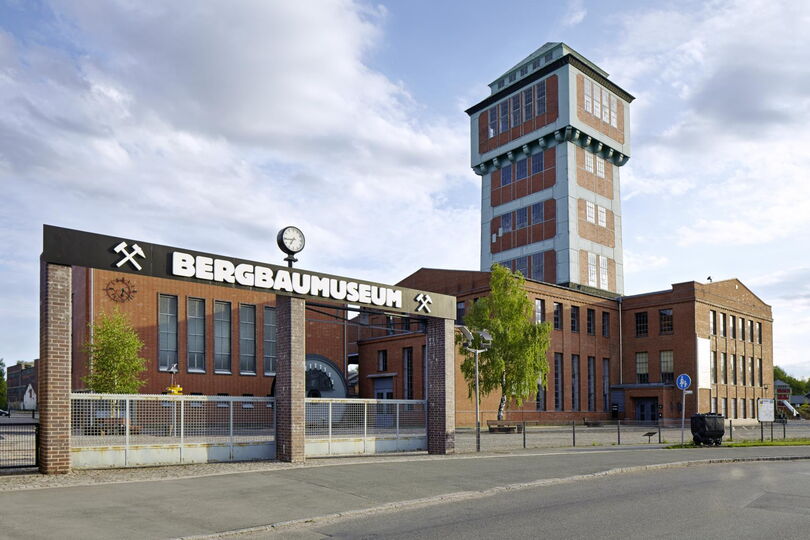 Das Bild zeigt den Eingangsbereich des Bergbaumuseums in Oelsnitz. Über dem Tor steht in großen weißen Buchstaben "Bergbaumuseum". Der Eingang ist von einem Metallzaun umgeben, und rechts im Bild sieht man das hohe Turmgebäude des Museums. Die Gebäude sind aus roten Ziegelsteinen gebaut, und der Himmel ist teilweise bewölkt.