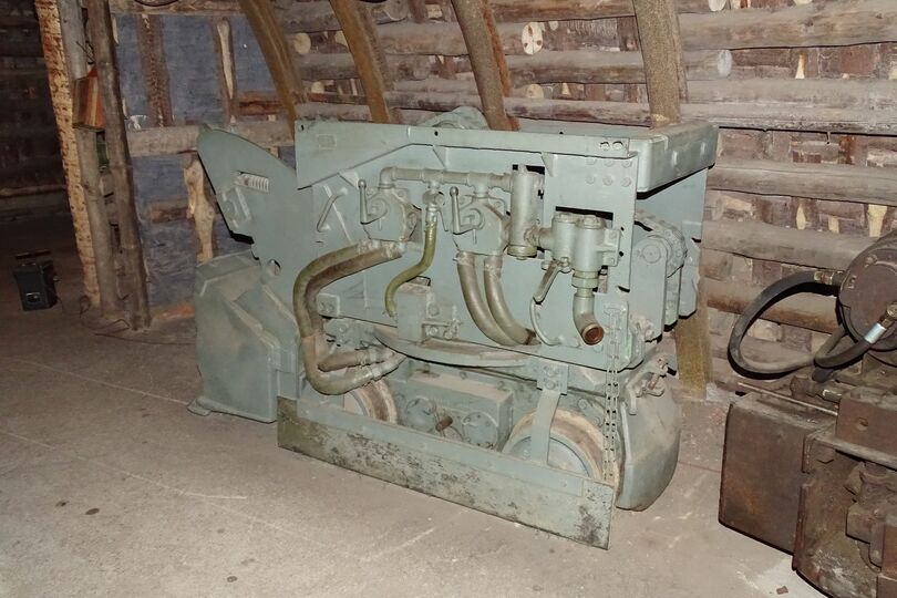 Es ist ein Wurfschauffellader aus Metall in einem Anschauungsbergwerk dargestellt. Das Gerät besteht aus einem Fahrgestell und einem Oberwagen. Es wird von vorne gezeigt. Dort befindet sich eine Schaufel, mit der man Gesteine aufnehmen konnte.