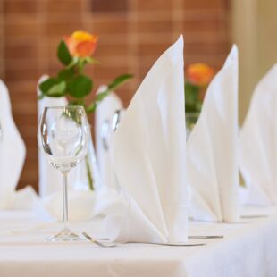 Ein detaillierter Blick auf eine festlich gedeckte Tafel. Weiße Servietten sind kunstvoll gefaltet und neben klaren Weingläsern platziert. In der Mitte des Tisches stehen kleine Vasen mit orangefarbenen Rosen. Der Hintergrund zeigt eine Backsteinwand.