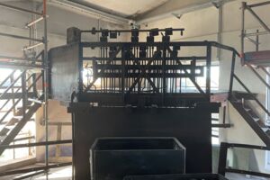 Dieses Bild zeigt einen Weichenstellerstand, der sich in einem Raum mit Gerüsten und Metallstrukturen befindet. Der Weichenstellerstand besteht aus einem schwarzen Metallgestell, auf dem verschiedene Hebel oder Mechanismen angebracht sind. Rechts und links führen Treppen aus Metall nach oben, und im Hintergrund sind Fenster sowie einige Baugerüste zu erkennen.