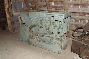 Es ist ein Wurfschauffellader aus Metall in einem Anschauungsbergwerk dargestellt. Das Gerät besteht aus einem Fahrgestell und einem Oberwagen. Es wird von vorne gezeigt. Dort befindet sich eine Schaufel, mit der man Gesteine aufnehmen konnte.