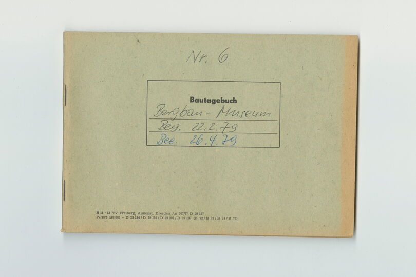 Die Abbildung zeigt ein grünbeiges Heft. Es ist im Querformat und ungefähr DIN A5 groß. Auf ihm befindet sich mittig ein schwarzer rechteckiger Rahmen. In diesem ist der Schriftzug „Bautagebuch“ gedruckt. Darunter sind Linien. Darauf steht handschriftlich: „Bergbau-Museum/Beg. 22.2.79/ Bee. 26.4.79“. Über dem Rahmen befindet sich der handschriftliche Vermerkt „Nr. 6“.  Links unten sind noch kleingedruckte Angaben zum Hersteller vorhanden.