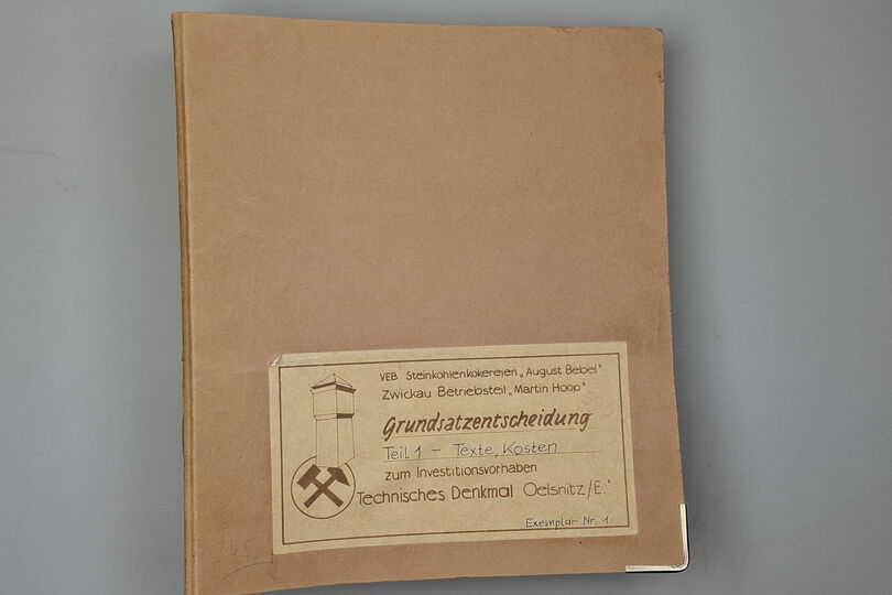 Es ist ein brauner Aktenordner abgebildet. Auf ihm befindet sich unterem Bereich ein Schild. Darauf sind links eine Umrisszeichnung des Förderturms und die Bergmannsinsignien Schlägel und Eisen zu sehen. Daneben befindet sich der Text: „VEB Steinkohlenkokerei „August Bebel“. Zwickauer Betriebsteil „Martin Hoop“. Grundsatzentscheidung. Teil 1 – Texte, Kosten. zum Investitionsvorhaben. Technisches Denkmal Oelsnitz/E. Exemplar Nr. 1“.