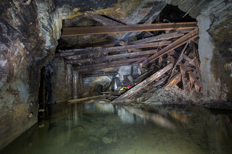 Das Bild zeigt einen Blick in ein Bergwerk. Im Fordergrund ist Wasser. Weiter hinten sieht man eingestürtzte Eisenträger. Ein Berarbeiter beichtigt den Einsturz.