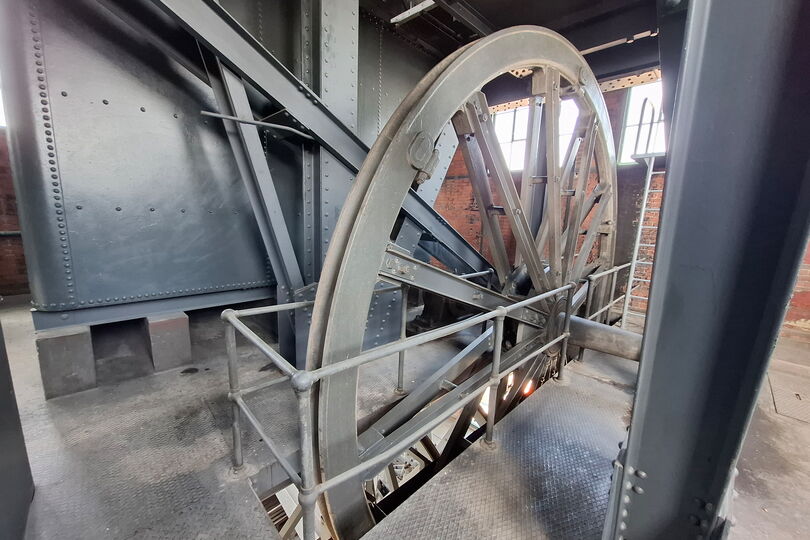 Es ist eine große Scheibe, die Leitscheibe, abgebildet. Sie ist jedoch nur ungefähr zur Hälfte sichtbar, während die andere Hälfte durch einen Schlitz im Boden geht. Die Metallscheibe ist nicht durchgehend gefüllt, sondern sie hat Speichen. Sie sieht damit so ähnlich aus wie das Rad eines Fuhrwerks aus Holz. Um die Scheibe herum sind Teile der Konstruktion des Turms abgebildet.