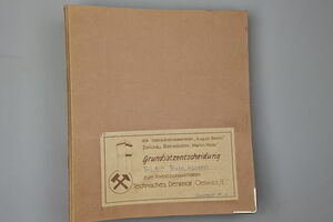Es ist ein brauner Aktenordner abgebildet. Auf ihm befindet sich unterem Bereich ein Schild. Darauf sind links eine Umrisszeichnung des Förderturms und die Bergmannsinsignien Schlägel und Eisen zu sehen. Daneben befindet sich der Text: „VEB Steinkohlenkokerei „August Bebel“. Zwickauer Betriebsteil „Martin Hoop“. Grundsatzentscheidung. Teil 1 – Texte, Kosten. zum Investitionsvorhaben. Technisches Denkmal Oelsnitz/E. Exemplar Nr. 1“.
