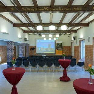 Der Raum ist für eine Präsentation oder Veranstaltung vorbereitet. Stuhlreihen sind auf den Bildschirm gerichtet, auf dem ein Bild von einem Bergbaugebäude zu sehen ist. Im Vordergrund stehen Tische mit roten Stofftüchern und Blumenvasen. Die Wände sind mit Backstein verkleidet, und die Decke hat sichtbare Holzbalken.