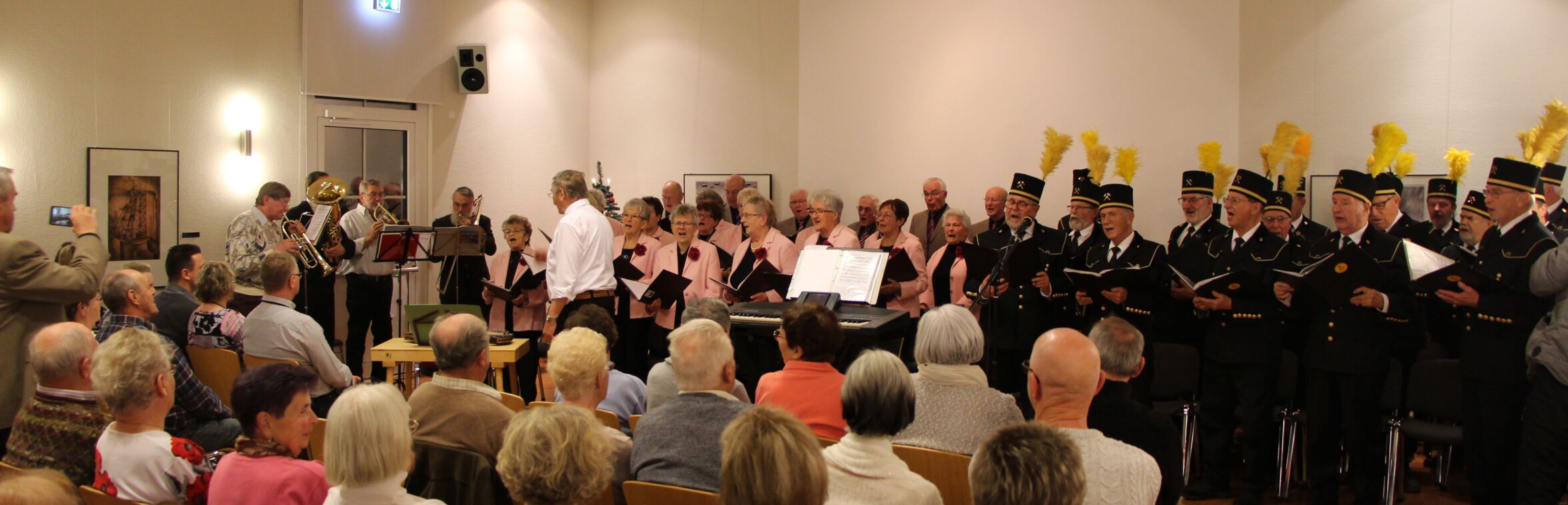 In einem Saal singen mehrere Männer in traditioneller Bergmannskleidung, die aus schwarzen Uniformen mit hohen Hüten und gelben Federn besteht. Im Hintergrund spielen Musiker Blasinstrumente, während das Publikum auf Stühlen sitzt und zuhört. Über den Anwesenden hängt ein roter Herrnhuther Stern von der Decke. Die Wände des Raumes sind hell, und die Atmosphäre wirkt festlich.