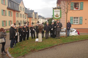 Eine Gruppe von Männern in schwarzer Bergmannsuniform steht um eine kleine Bronzeskulptur auf einem Sockel. Einer der Männer hält eine große Fahne mit der Aufschrift „Förderverein Bergbaumuseum Oelsnitz/Erzgebirge e.V.“. Im Hintergrund sind mehrere pastellfarbene Gebäude mit grünen Fensterläden zu sehen. Links im Bild fotografiert eine Frau die Szene, während zwei Personen aus einem der oberen Fenster des Gebäudes zuschauen.