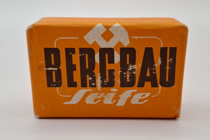 Es ist ein Stück Seife abgebildet. Dieses hat die Form eines Quaders und ist verpackt. Die Verpackung ist orange. Auf ihr befindet sich der Aufdruck „Bergbau“ in schwarzer Farbe und Großbuchstaben. Darüber und teilweise dahinter sind die Bergmannsinsignien, Schlägel und Eisen, abgebildet. Unter dem Aufdruck steht der weiße Schriftzug „Seife“. Die Verpackung ist an ein paar Stellen etwas beschädigt.