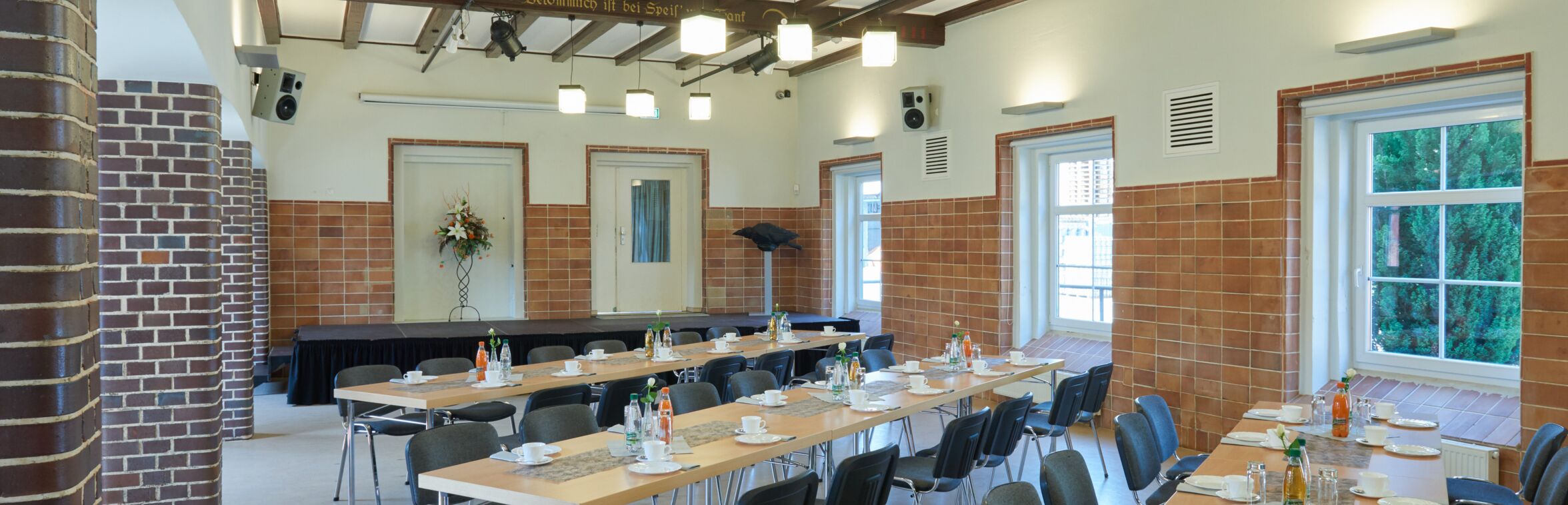 Ein rechteckiger Raum mit gedeckten, rechteckigen Tischen und Stühlen. Auf den Tischen stehen Wasserflaschen, Getränkeflaschen und Geschirr. Die Decke des Raumes ist mit Holzbalken verziert, auf denen der Text „Ein fröhlicher Scherz, ein lustiger Schwank, bekommlich ist bei Speis' und Trank“ in goldener Schrift steht. Im Hintergrund befindet sich eine kleine Bühne mit einem Blumenarrangement auf einem Ständer.