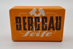 Es ist ein Stück Seife abgebildet. Dieses hat die Form eines Quaders und ist verpackt. Die Verpackung ist orange. Auf ihr befindet sich der Aufdruck „Bergbau“ in schwarzer Farbe und Großbuchstaben. Darüber und teilweise dahinter sind die Bergmannsinsignien, Schlägel und Eisen, abgebildet. Unter dem Aufdruck steht der weiße Schriftzug „Seife“. Die Verpackung ist an ein paar Stellen etwas beschädigt.