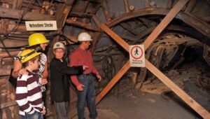 Das Bild zeigt eine Frau im schwarzen Hemd, die eine Familie durch einen alten Stollen im Anschauungsbergwerk Oelsnitz führt. Die Familie trägt Helme und steht vor einer gesperrten Strecke, die mit einem Schild „Fahrung verboten!“ markiert ist. Die Frau zeigt auf die verbrochene Strecke und erklärt vermutlich etwas über den Stollen. Die Wände des Stollens bestehen aus Holz, und die Beleuchtung sorgt für eine authentische, gedämpfte Bergwerksatmosphäre.