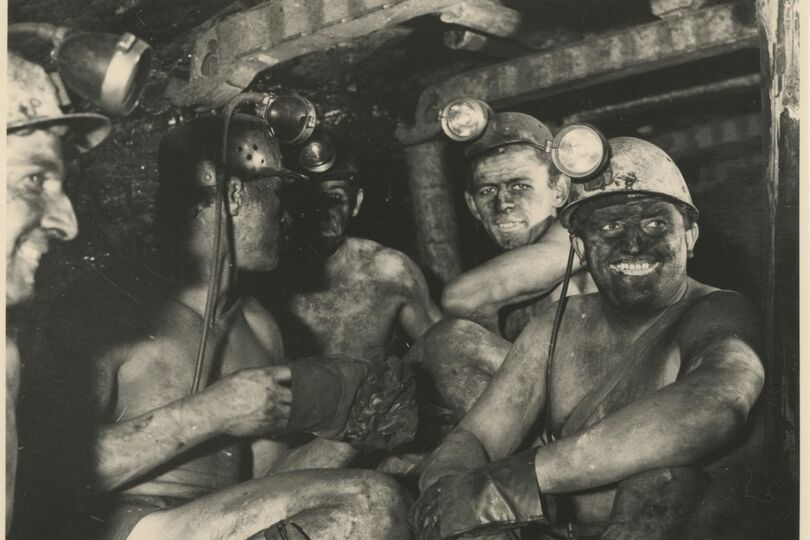 Es handelt sich um eine Schwarz-Weiß-Fotografie. Auf dieser sind fünf Männer abgebildet. Sie sitzen in einem Abbauort unter Tage und schauen zum Teil in die Kamera. Ihre Gesichter und ihre nackten Oberkörper sind von Kohlenstaub verschmutzt. Die Bergleute haben Schutzhelme mit aufgesteckten Lampen auf.