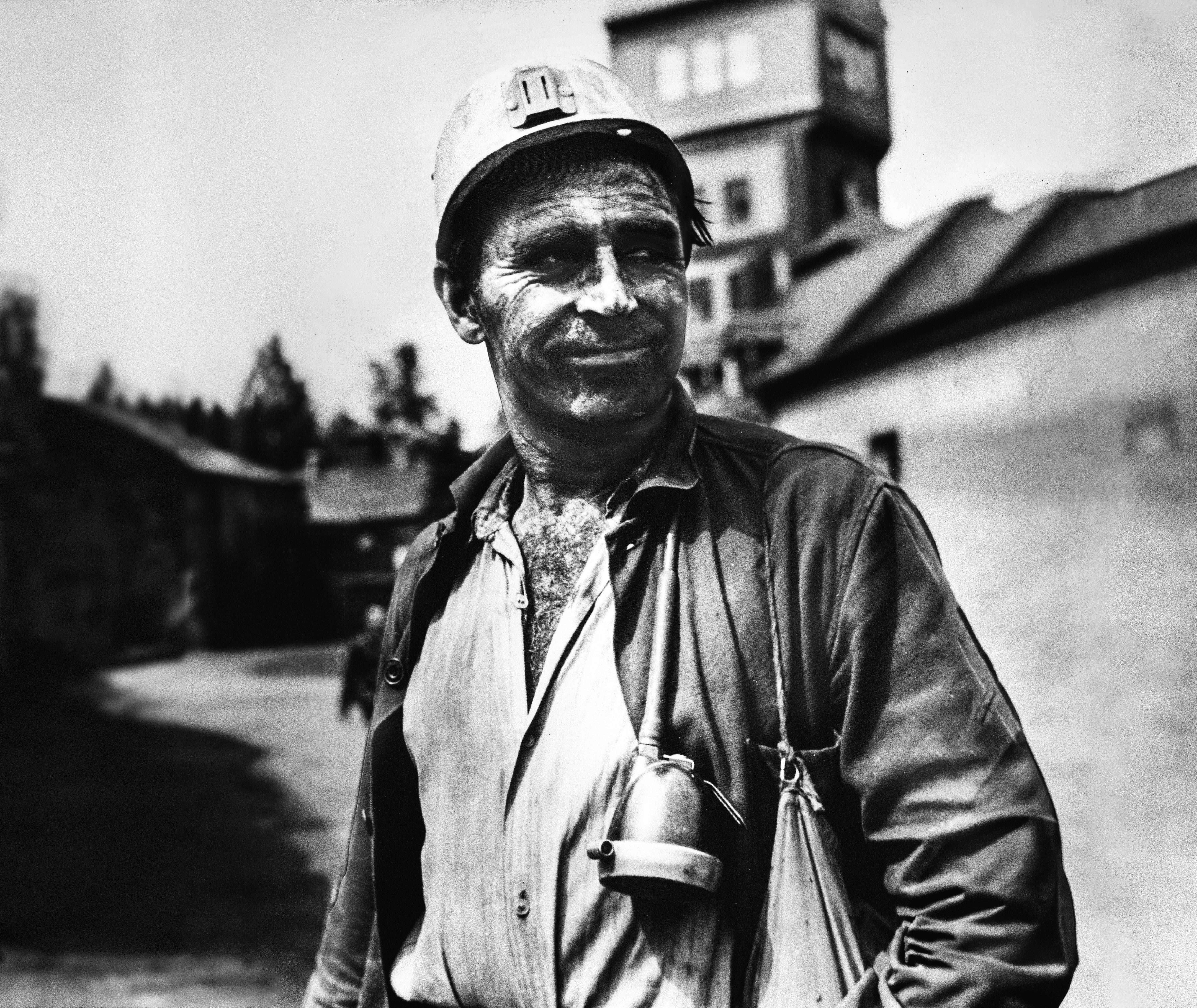 Ein Bergmann steht im Freien, trägt einen Schutzhelm mit einer Lampe und ist von Kohlenstaub bedeckt. Seine Arbeitskleidung ist geöffnet, und er hat eine Lampe an seiner Brust befestigt. Im Hintergrund sind unscharf Industriegebäude des Bergbaus im Lugau-Oelsnitzer Revier zu sehen.
